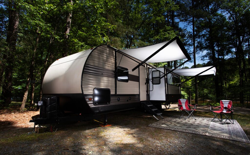 White RV with awnings out, 2 red chairs on top of a rug, all on a forest campground.