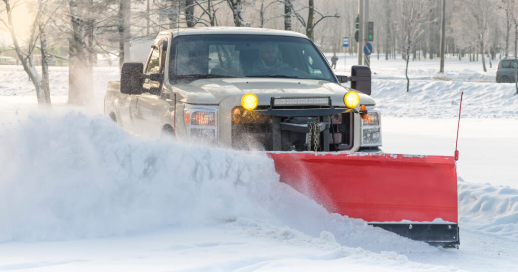 Picture of a white truck with a red snow plow plowing a side street during the day.