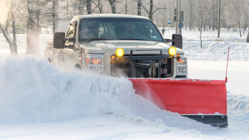 Picture of a white truck with a red snow plow plowing a side street during the day.