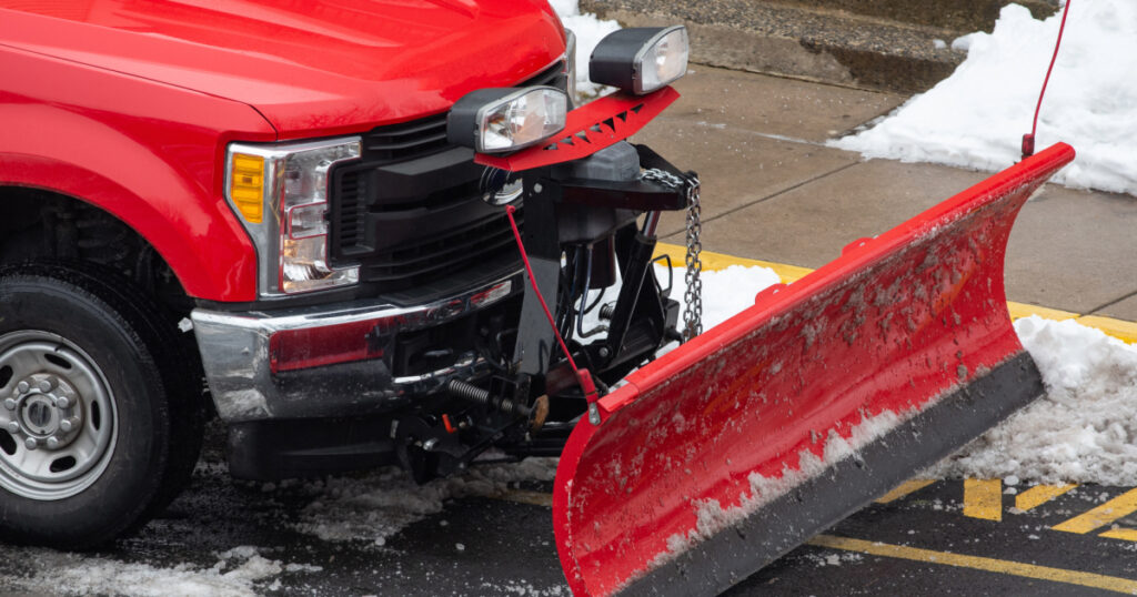 Side view of a red snow plow attached to a red truck parked on the side of a snowy street.