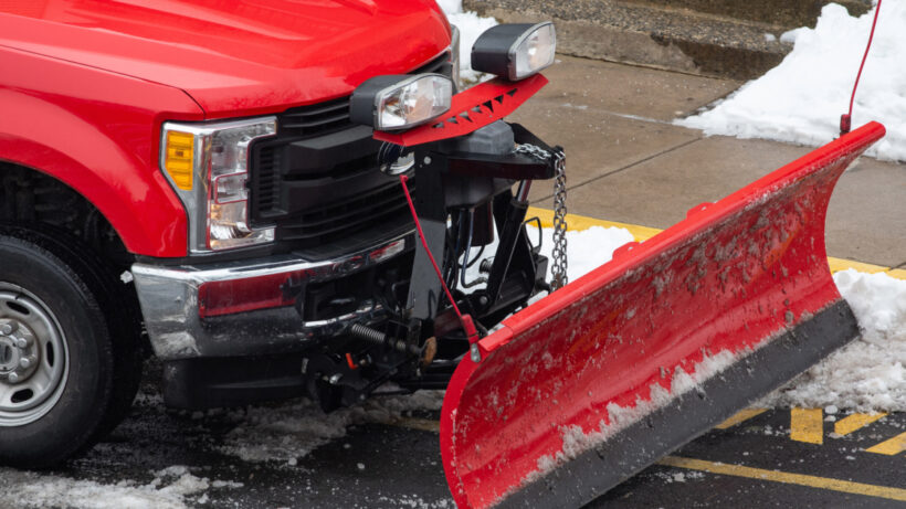 Side view of a red snow plow attached to a red truck parked on the side of a snowy street.