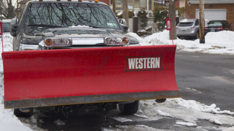 Front facing view of a Western snow plow mounted on a truck on a snowy street.
