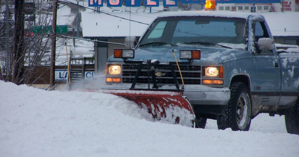 Blue old school truck with snow plow and headlight attached to the front bumper, plowing snow in the street.