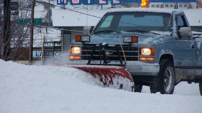 Blue old school truck with snow plow and headlight attached to the front bumper, plowing snow in the street.